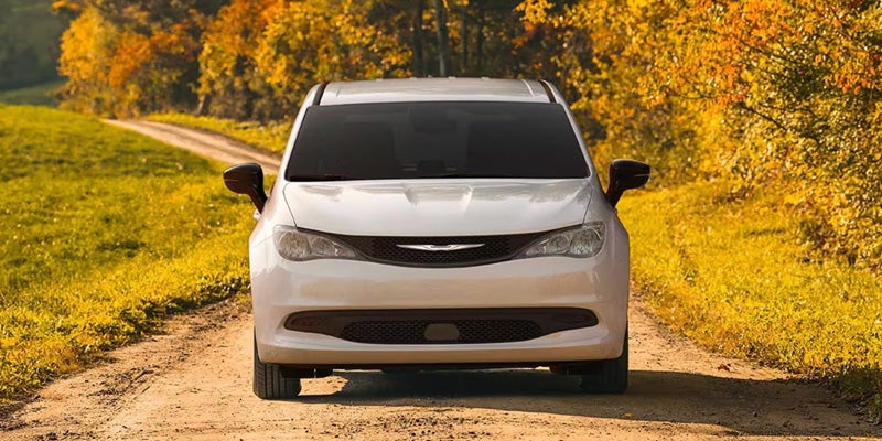 Front view of a white minivan parked on a dirt road, surrounded by fields and trees with yellow-orange fall foliage