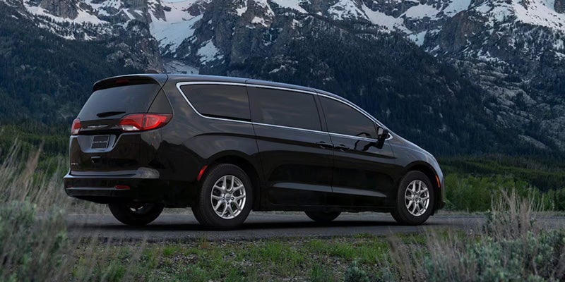 Rear view of a black Chrysler Pacifica minivan parked on a road with snowy mountains and pine trees in the background