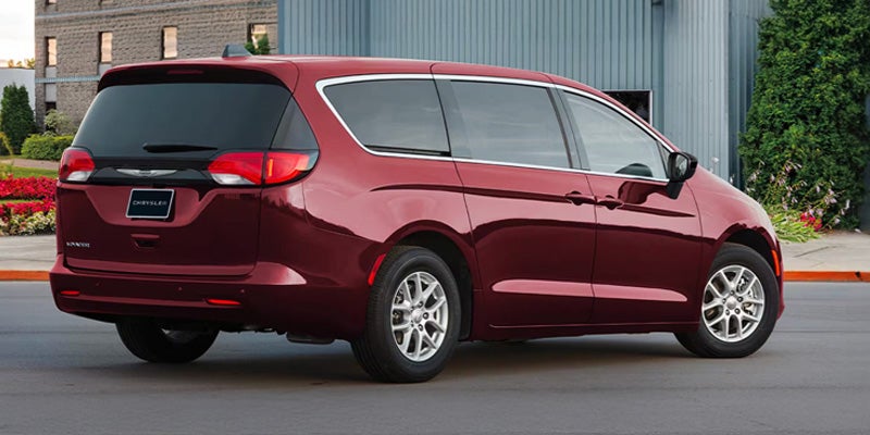 Rear 3/4 view of a dark red Chrysler Pacifica minivan parked on asphalt with a building and plants in the background