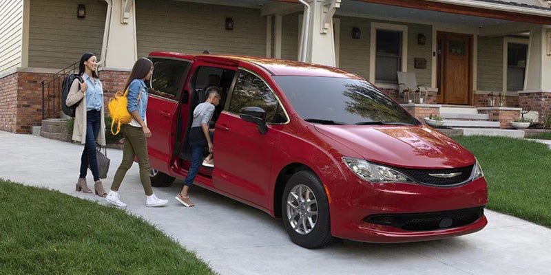Two women and a child load into a red minivan on a driveway in front of a house