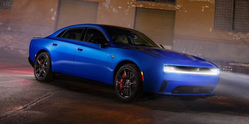 A bright blue 2024 Dodge Charger parked with its headlights on, angled slightly right against a dimly lit building