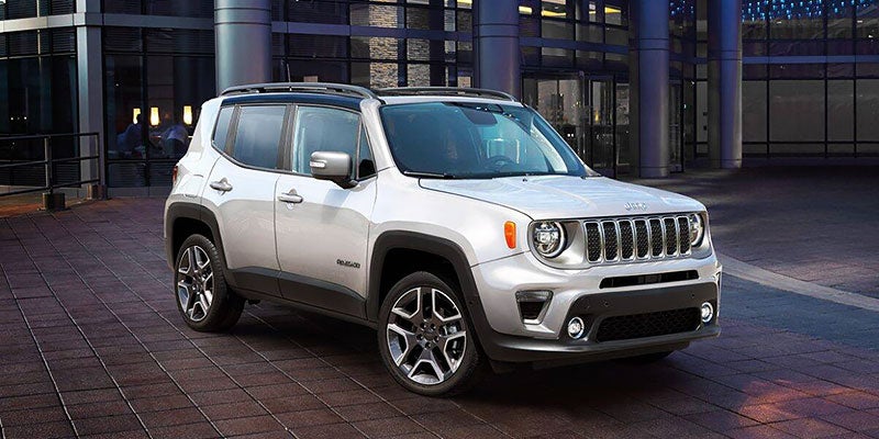 A light gray Jeep Renegade with a black roof and roof rack is parked on dark brown cobblestones in front of a modern glass building