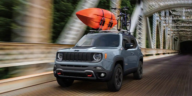 Gray Jeep Renegade driving on a bridge, equipped with a roof rack holding an orange kayak and a bike