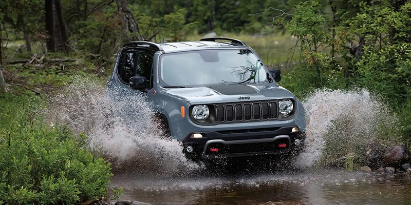 A gray Jeep Renegade splashing through a shallow river in a lush green forest