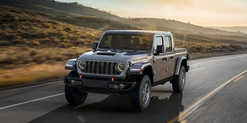 Silver four-door pickup truck driving on a paved road through a hilly countryside at sunset