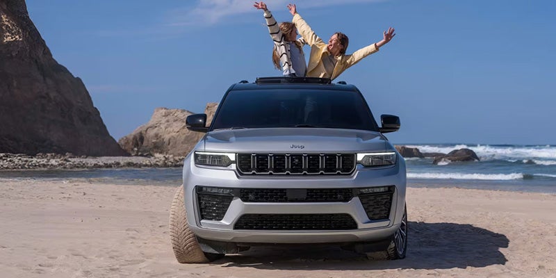 Silver Jeep SUV on a sandy beach with two people waving out of the sunroof
