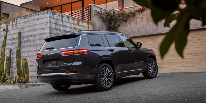 Gray Jeep Grand Cherokee L, seen from the rear passenger side, parked on an asphalt driveway near a stone building