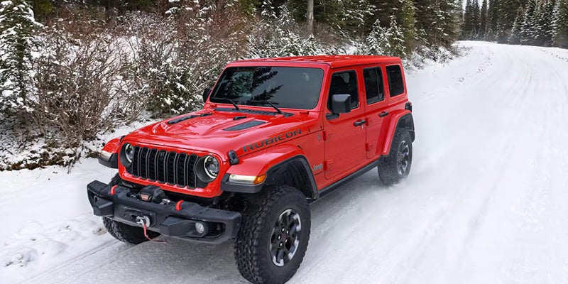 A vibrant red Jeep Wrangler Rubicon driving on a snow-covered road lined with snowy evergreen trees