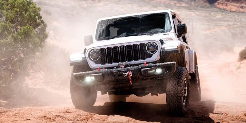 A white Jeep Wrangler Rubicon kicks up orange dust while driving off-road through rugged terrain
