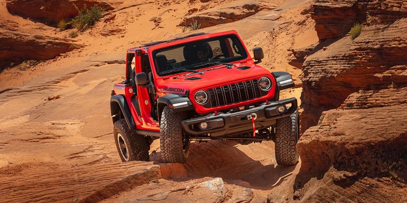 A bright red Jeep Wrangler Rubicon off-roading on a sandy, rocky path with its left front wheel elevated