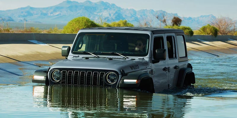 A dark-colored four-door SUV drives through shallow water in a concrete drainage canal, with water reaching the lower section of the vehicle. Mountain