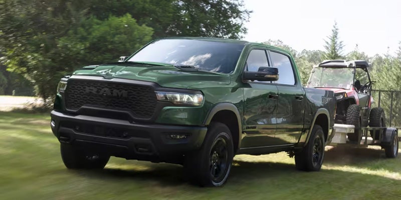 A dark green Ram pickup truck drives left on green grass, towing a red off-road vehicle on a utility trailer