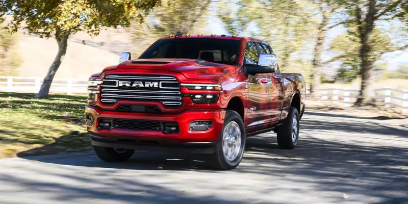 Front view of a shiny red Ram 3500 heavy-duty pickup truck moving on a paved road with trees and a white fence.