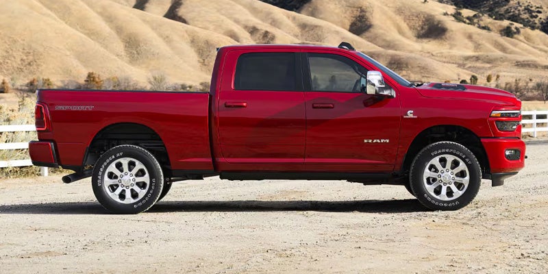 Side view of a red Dodge Ram pickup truck with black tinted windows on a dirt road with rolling hills