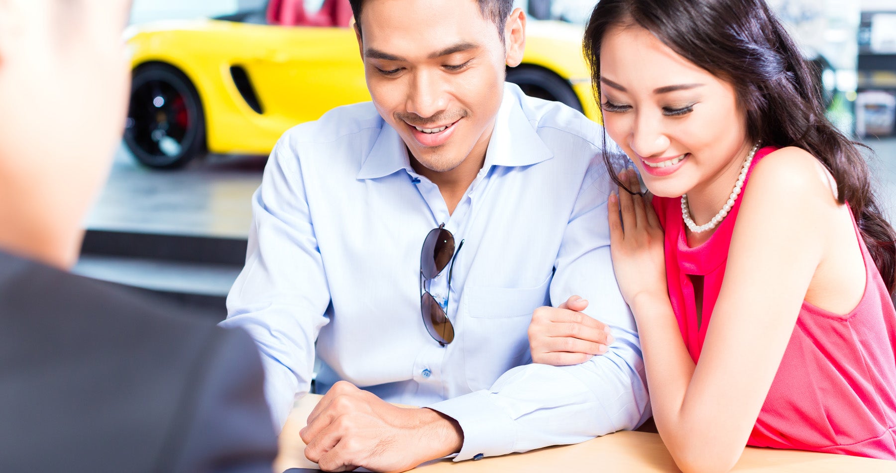 A smiling couple, dressed casually, looking down at a table while a person facing away from the camera talks to them