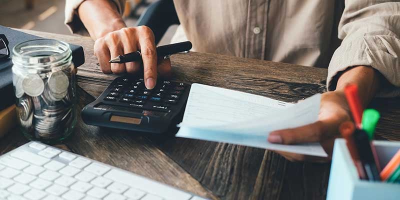 Person at a wooden desk manually calculating finances with a calculator, pen, and paperwork