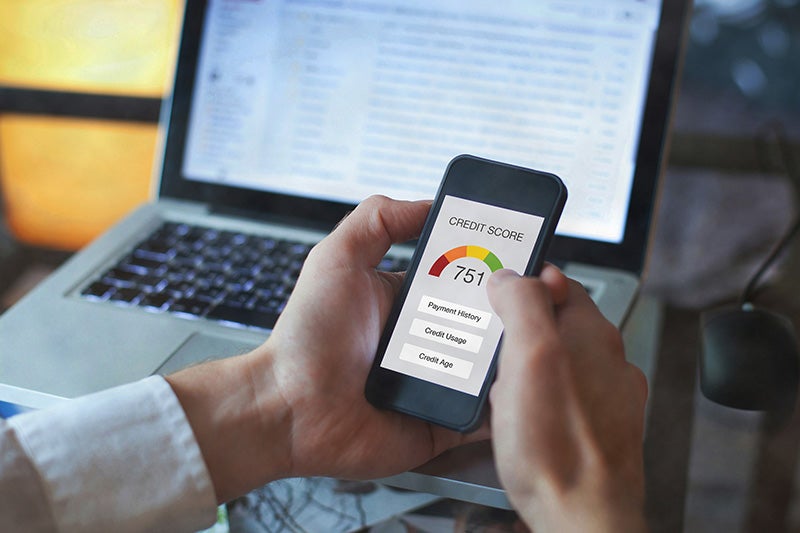 Hands holding a smartphone displaying a credit score of 751 on an app, with a laptop in the background