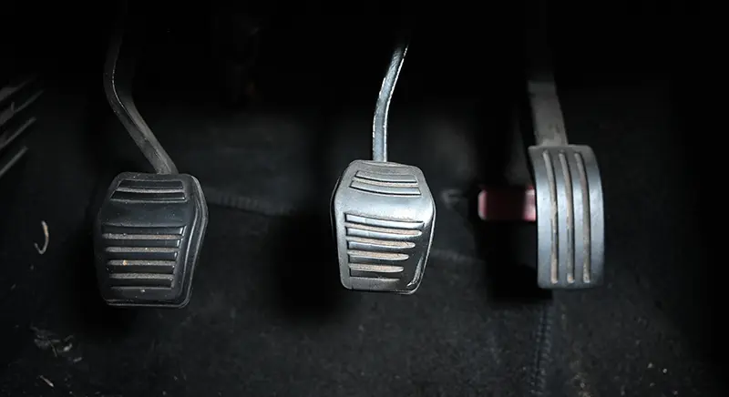 Close-up view of three vehicle pedals in a car footwell, showing the clutch, brake, and accelerator pedals mounted above a dark floor mat