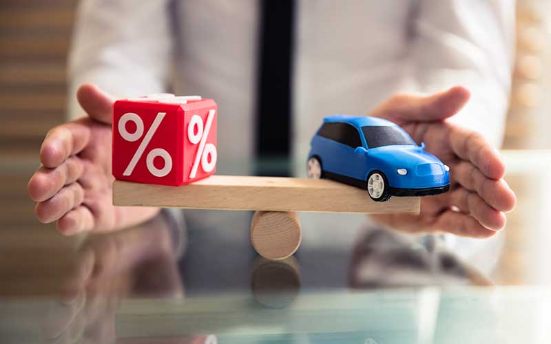 Hands balancing a blue toy car and a red cube with percentage symbols on a wooden seesaw, suggesting car interest rates