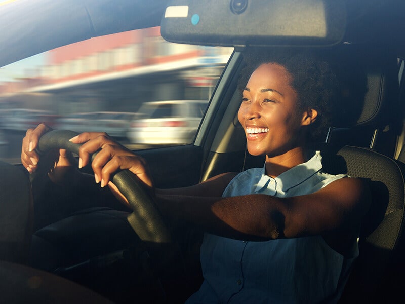 A smiling woman with natural hair and light blue top, driving a car with both hands on the steering wheel