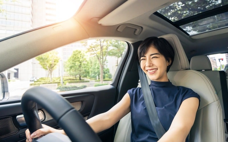 A smiling woman with short dark hair in a navy shirt and seatbelt, driving a car with a sunroof