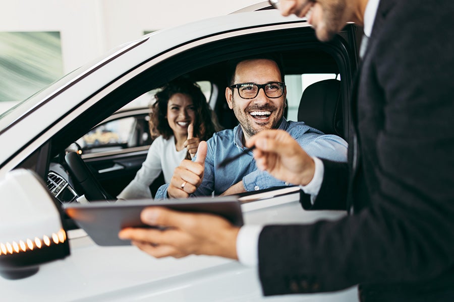 Two happy customers in a white car, giving thumbs up, talking to a salesperson with a tablet