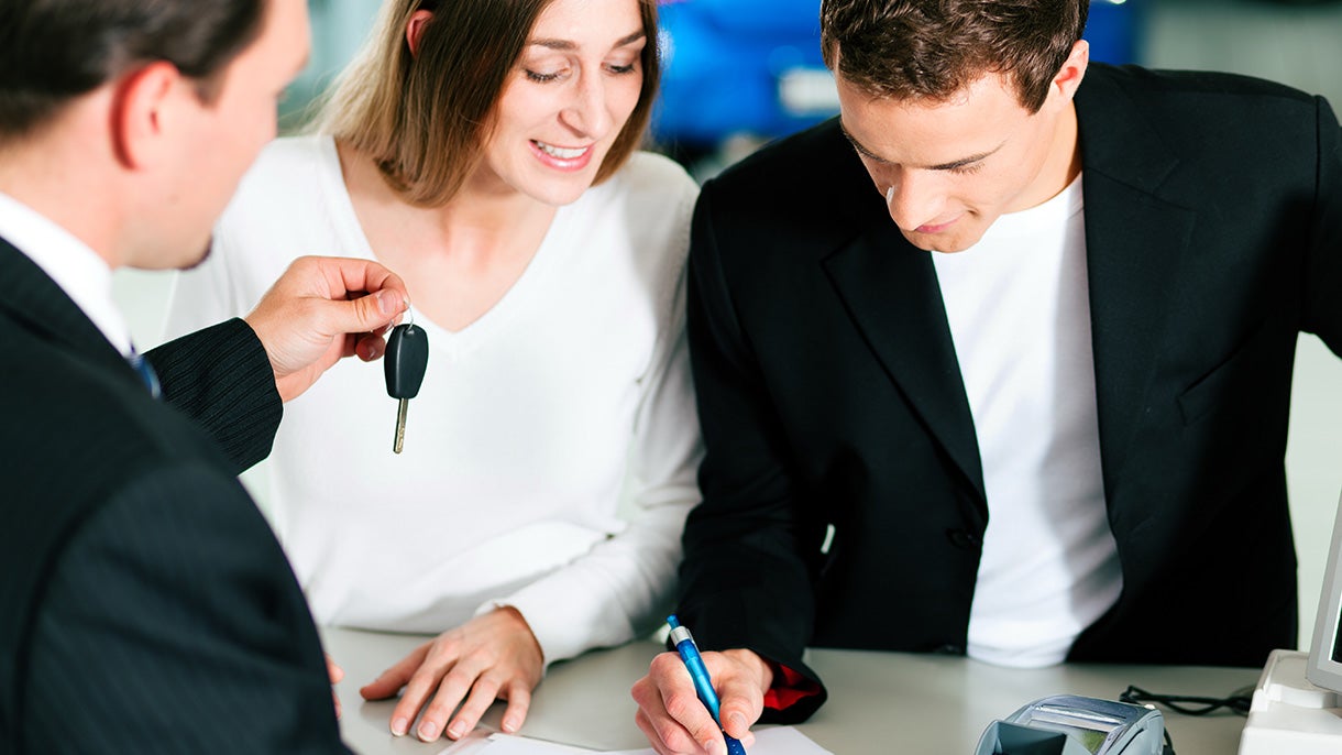 A car salesman handing keys to a smiling couple, the man signing papers for a new car
