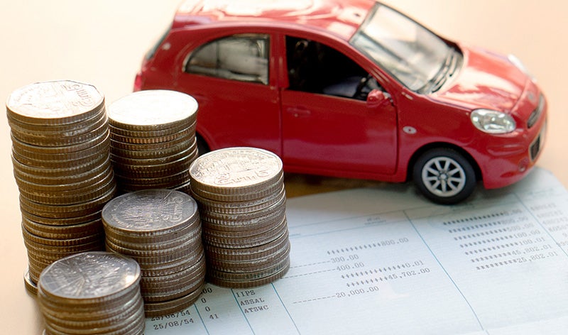 A red toy car sits on a bank book, surrounded by stacks of coins on a light-colored surface
