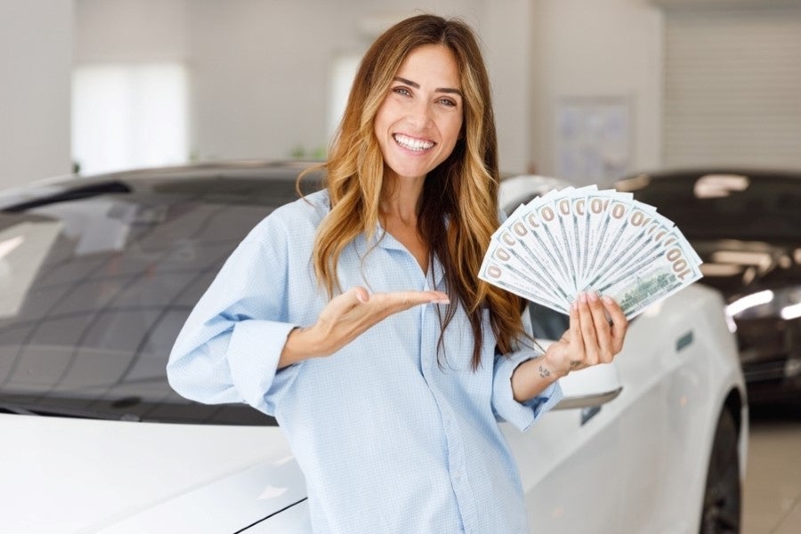 A smiling woman stands in front of a white car, holding a fan of hundred-dollar bills