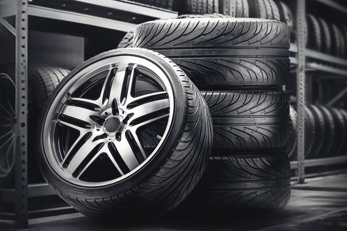 A stack of black performance tires in a warehouse or tire storage area, with a silver alloy wheel leaning against the front.