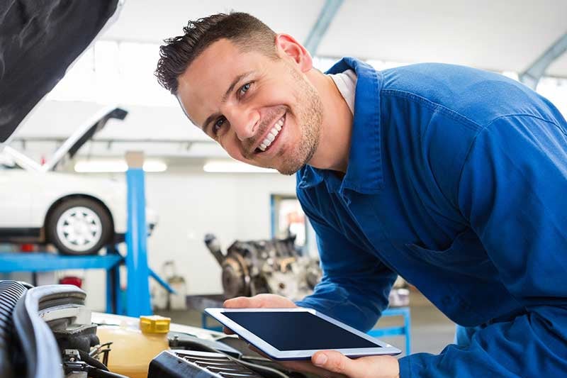 Smiling mechanic in blue coveralls holding a tablet and leaning over a car engine, with a car lift in the background