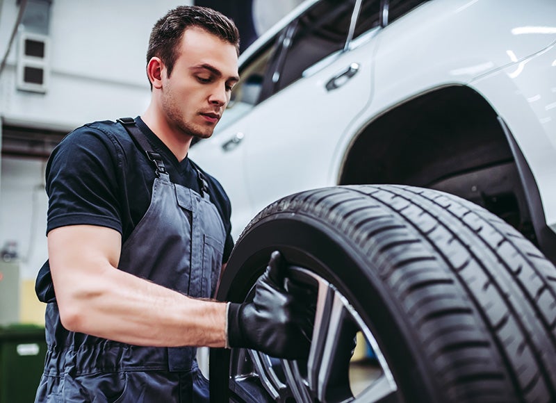 A young male mechanic in a gray overall preparing to install a tire onto a white car