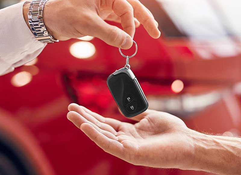 A man's hand, wearing a watch, hands a black car key fob to an outstretched hand, against a red car