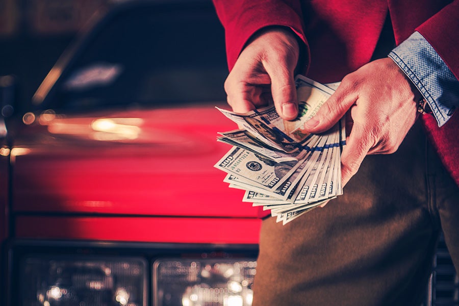 Close-up of a person holding and counting several U.S. dollar bills in front of a red car