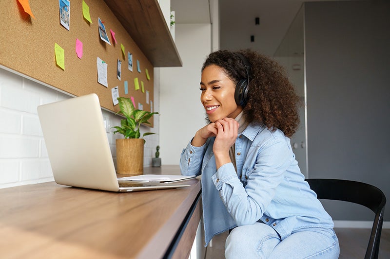 Smiling young Black woman with curly hair and headphones looking at a laptop computer